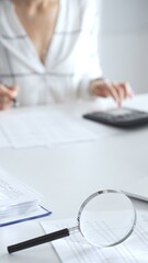 Magnifying glass on financial documents, with an accountant using a calculator and taking notes, emphasizing accounting, finance, and tax preparation, close up vertical view. Audit and taxes