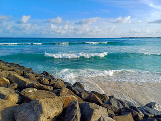 Rugged coastline on a sunny day, dominated by a collection of large, weathered rocks, with gentle, frothy waves rolling onto the shore from the turquoise-colored water. Related to ocean scenery
