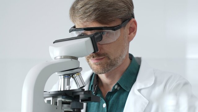 Senior man scientist wearing protective glasses and lab coat analyzing samples with a microscope in laboratory setting. Medicine, health care and science concept - Powered by Adobe