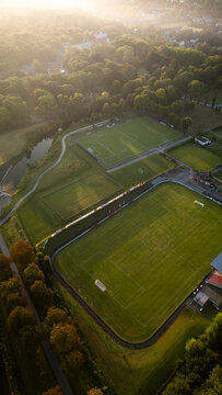 Vertical aerial top-down view of sports fields and running track surrounded by green grass and autumn-colored trees at sunrise.