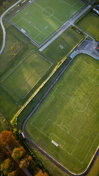 Vertical aerial top-down view of sports fields and running track surrounded by green grass and autumn-colored trees at sunrise.