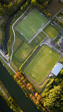 Vertical aerial top-down view of sports fields and running track surrounded by green grass and autumn-colored trees at sunrise.