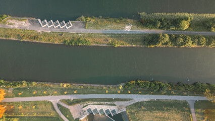 Aerial view of a canal with parallel paths, green vegetation and water obstacles, taken with a drone in warm morning light.
