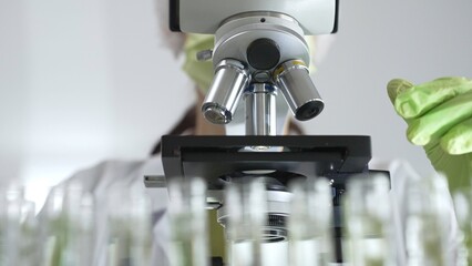 Scientist in green protective mask and gloves holding test tube with plant sample, researching botany and medical applications using a microscope. Medicine, healthcare and science concept