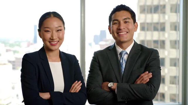 Two smiling professionals stand side by side, radiating confidence and success, in front of a bright window with a city view.