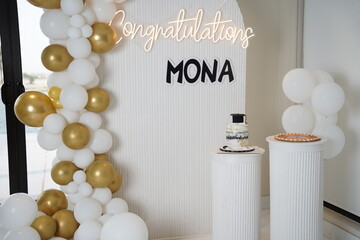 
Stylish celebration setup featuring a white and gold balloon arch, decorative backdrop with &ldquo;Congratulations Mona&rdquo; sign, and cakes on white columns, perfect for graduation or special occasions
