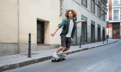 Young curly haired young man balancing on a longboard while cruising downhill on a paved road in an urban setting
