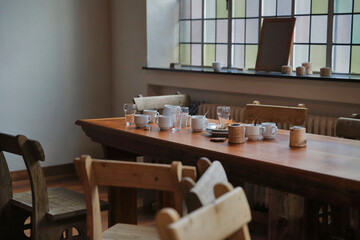 Rustic wooden dining table with ceramic cups plates and glassware arranged in traditional interior with stained glass window background