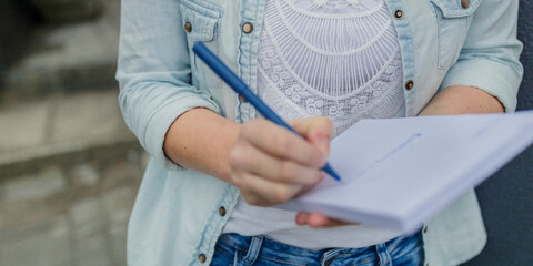 Person in denim jacket and lace top writing on paper with blue pen in casual outdoor setting