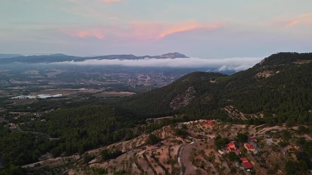 Drone video of a mountain valley in Spain during sunset, with low clouds stretching along the horizon and scattered houses among the forested hills.