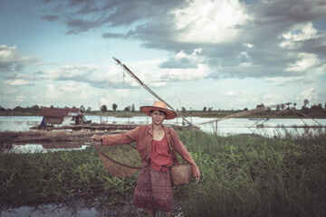 Woman in Traditional Clothing Standing in Wetland with Fishing Equipment and Scenic Background Under Dramatic Sky