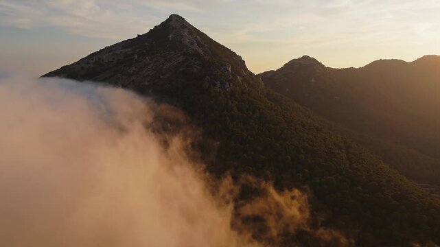 Drone video over Spanish mountain peak covered with forest, with golden sunset light illuminating drifting clouds in the foreground.