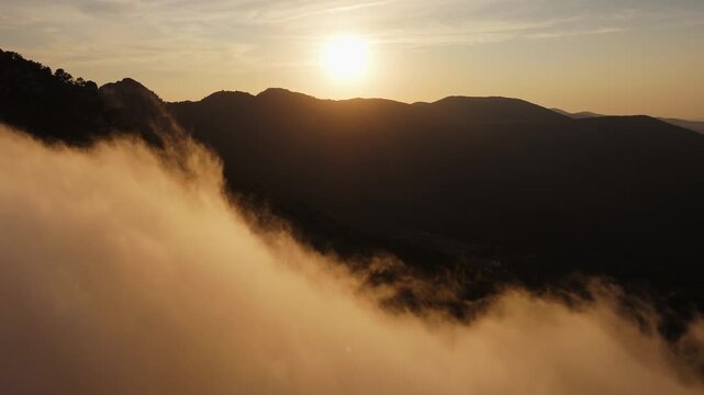 Drone video over Spanish mountain peak covered with forest, with golden sunset light illuminating drifting clouds in the foreground.