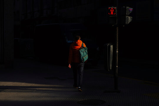 Person with a turquoise backpack walks through a dark street under dramatic lighting, passing a red pedestrian signal, creating a cinematic urban atmosphere.