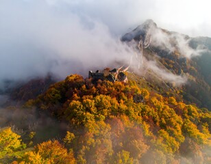 Fototapeta premium Aerial view of a medieval castle ruin perched atop a vibrant autumn-colored, forested mountain peak, shrouded in mist and clouds.