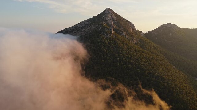 Drone video over Spanish mountain peak covered with forest, with golden sunset light illuminating drifting clouds in the foreground.