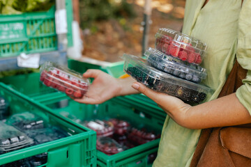 Close-up of hand with fresh berry in plastic packaging at local urban market. Organic produce on...