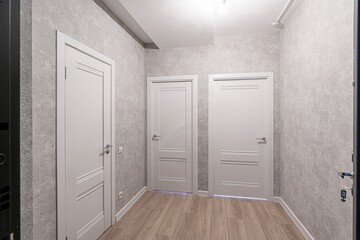 hallway with three white doors, gray textured walls, and light wood-like flooring. Bright overhead lighting illuminates the space