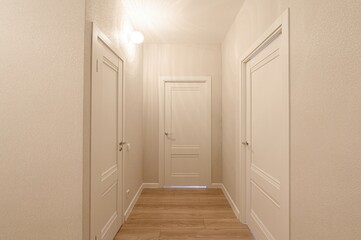 bright hallway with three white doors, wooden flooring, and textured walls. A round light fixture is visible on the ceiling. The scene is clean and minimalist