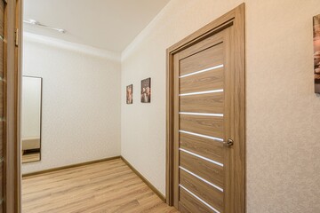 modern hallway with wooden flooring, light beige wallpaper, and a stylish wooden door with white stripes. Pictures hang on the wall. A mirror is visible through an open doorway