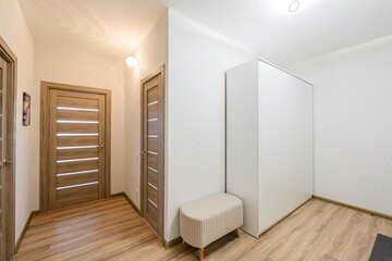 Bright hallway with wooden doors, a white wardrobe, and a small bench. The walls have a subtle pattern, and the floor is wooden