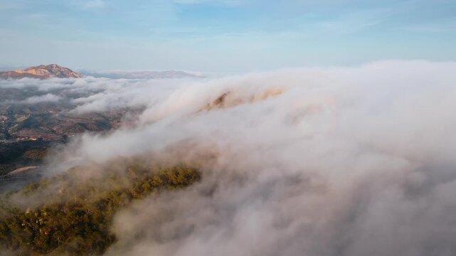 Drone video of Spanish mountains with dense clouds flowing over forested slopes, revealing peaks under clear blue sky.