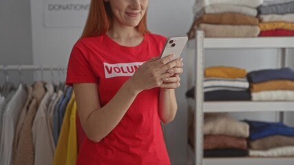 Woman wearing red volunteer shirt using smartphone in donation center fills room with textiles, embodying youth, charity spirit, focused indoors.