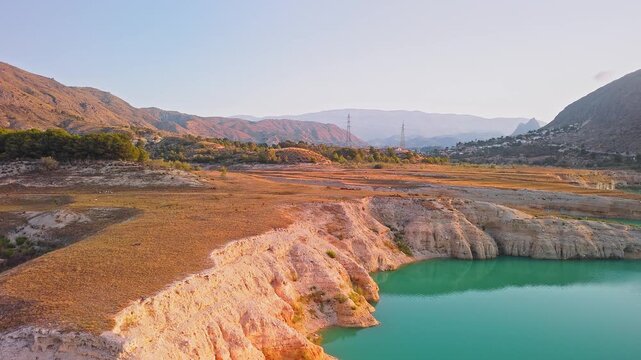 Drone video showing a sharp rocky peninsula surrounded by vibrant turquoise waters of a Spanish lake, bathed in warm golden hour sunlight.