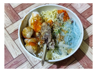 Bakso. A bowl of rib meatballs with yellow noodles, blue vermicelli, shredded cabbage, fried onions and chili sauce on a wooden patterned table. The meatballs look very delicious and delicious.