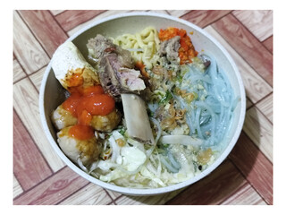 Bakso. A bowl of rib meatballs with yellow noodles, blue vermicelli, shredded cabbage, fried onions and chili sauce on a wooden patterned table. The meatballs look very delicious and delicious.