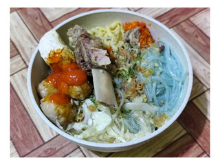 Bakso. A bowl of rib meatballs with yellow noodles, blue vermicelli, shredded cabbage, fried onions and chili sauce on a wooden patterned table. The meatballs look very delicious and delicious.