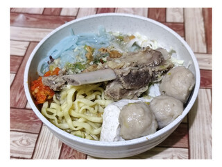 Bakso. A bowl of rib meatballs with yellow noodles, blue vermicelli, shredded cabbage, fried onions and chili sauce on a wooden patterned table. The meatballs look very delicious and delicious.