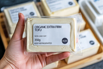 Hand holding firm tofu block in front of supermarket refrigerator. Concept of plant-based protein and vegan grocery shopping