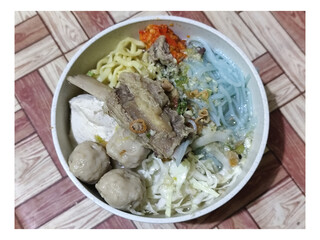 Bakso. A bowl of rib meatballs with yellow noodles, blue vermicelli, shredded cabbage, fried onions and chili sauce on a wooden patterned table. The meatballs look very delicious and delicious.