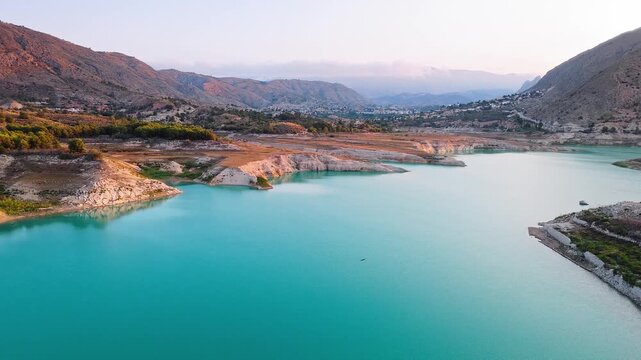 Drone video showing a vivid turquoise lake in Spain with rocky, sunlit shoreline and earthy cliffs creating striking contrast between water and land.