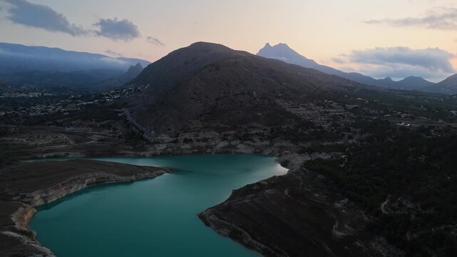 Drone video of a mountain lake in Spain at sunset, with calm turquoise water, rugged hills, and a dramatic sky blending warm and cool tones.