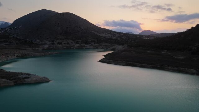 Drone video of a mountain lake in Spain at sunset, with calm turquoise water, rugged hills, and a dramatic sky blending warm and cool tones.