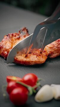 Chef cuts juicy grilled pork steak with sharp knife, garnished with fresh cherry tomatoes and garlic cloves on dark surface.