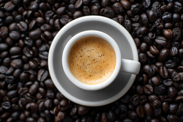 Obraz premium A top-down perspective of a tiny white espresso cup with frothy cream, resting on a saucer amidst a background of dark roasted coffee beans.