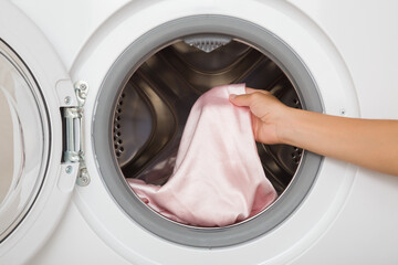 Young adult woman hands holding and unloading pink silk fabric from opened white washing machine. Clothes care. Closeup. Front view.