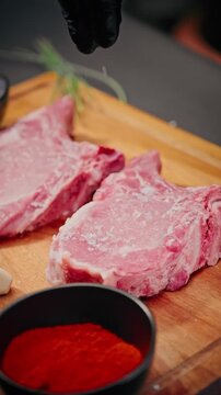 Close-up of chef sprinkling coarse salt on raw pork chops placed on a wooden board with paprika, garlic, and herbs for cooking preparation.