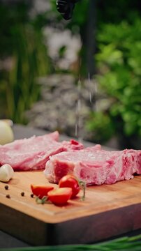 Chef&rsquo;s gloved hand sprinkles salt over raw pork ribs on wooden board with tomatoes, garlic, and peppercorns, preparing meat for cooking.