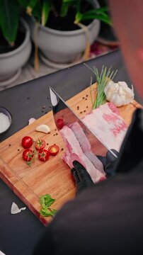Cook wearing black gloves slices raw pork ribs on wooden board surrounded by cherry tomatoes, garlic, chives, peppercorns, and fresh herbs.