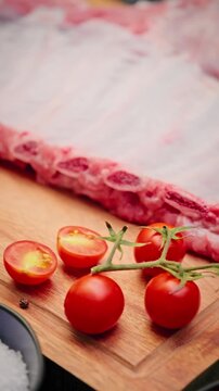 Close-up view of raw pork ribs on wooden board with vine tomatoes, garlic, paprika powder, and fresh herbs prepared for cooking.