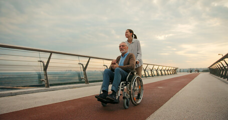 Woman pushes elderly man in wheelchair on bridge under cloudy sky