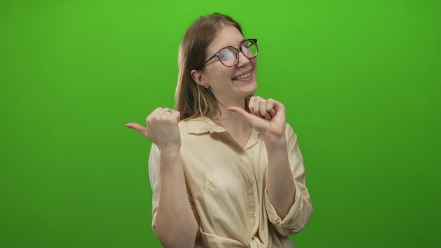 Smiling woman pointing both thumbs at her chest in green studio setting with eyeglasses and beige blouse; pride.