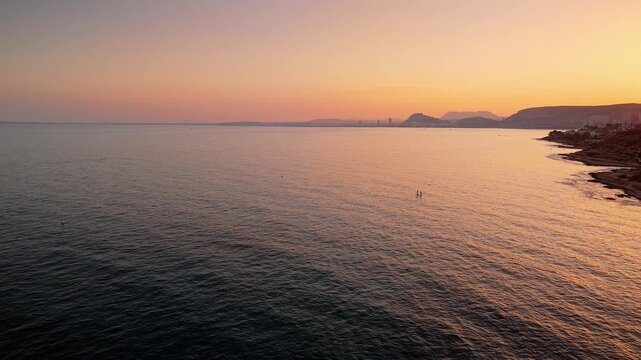 Drone video capturing Spanish coastline and city skyline at sunset, with golden light reflecting on calm sea and rugged shore.