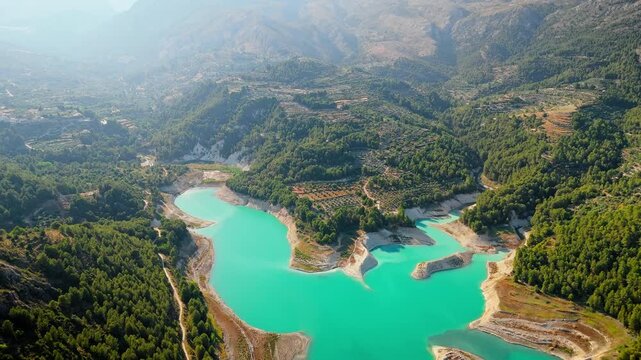 Drone video of the turquoise Guadalest Reservoir in Spain, surrounded by green hills and mountains, showing vivid water colors and scenic Mediterranean landscapes.