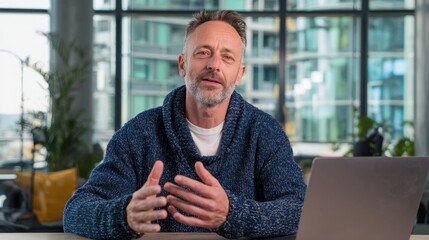 A mature man with a beard sits at a desk with a laptop, gesturing as he speaks, possibly during a video call or online presentation in a bright, modern setting.