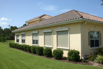 Exterior view of a modern suburban house with tiled roof, stucco walls, roller shutters on windows, and neat landscaping under a blue sky